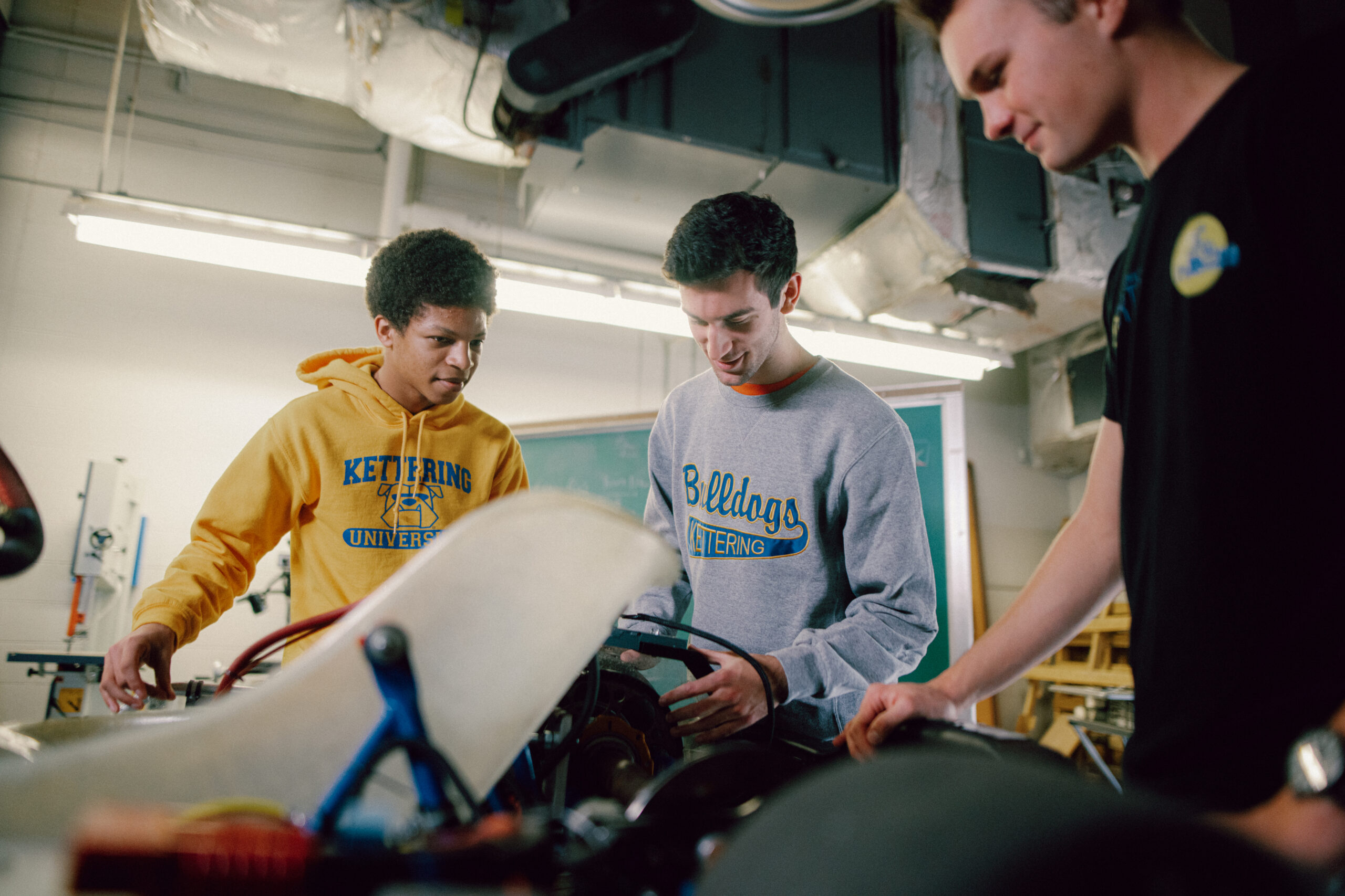 Three students are engaged in a hands-on project in a workshop. One student is wearing a yellow Kettering University hoodie, another is in a gray Bulldogs Kettering sweatshirt, and the third is in a black t-shirt. They are working together on what appears to be a mechanical or engineering project, likely involving vehicle components. The setting is a typical workshop with tools and equipment visible in the background, and the students are focused and collaborating on their task.