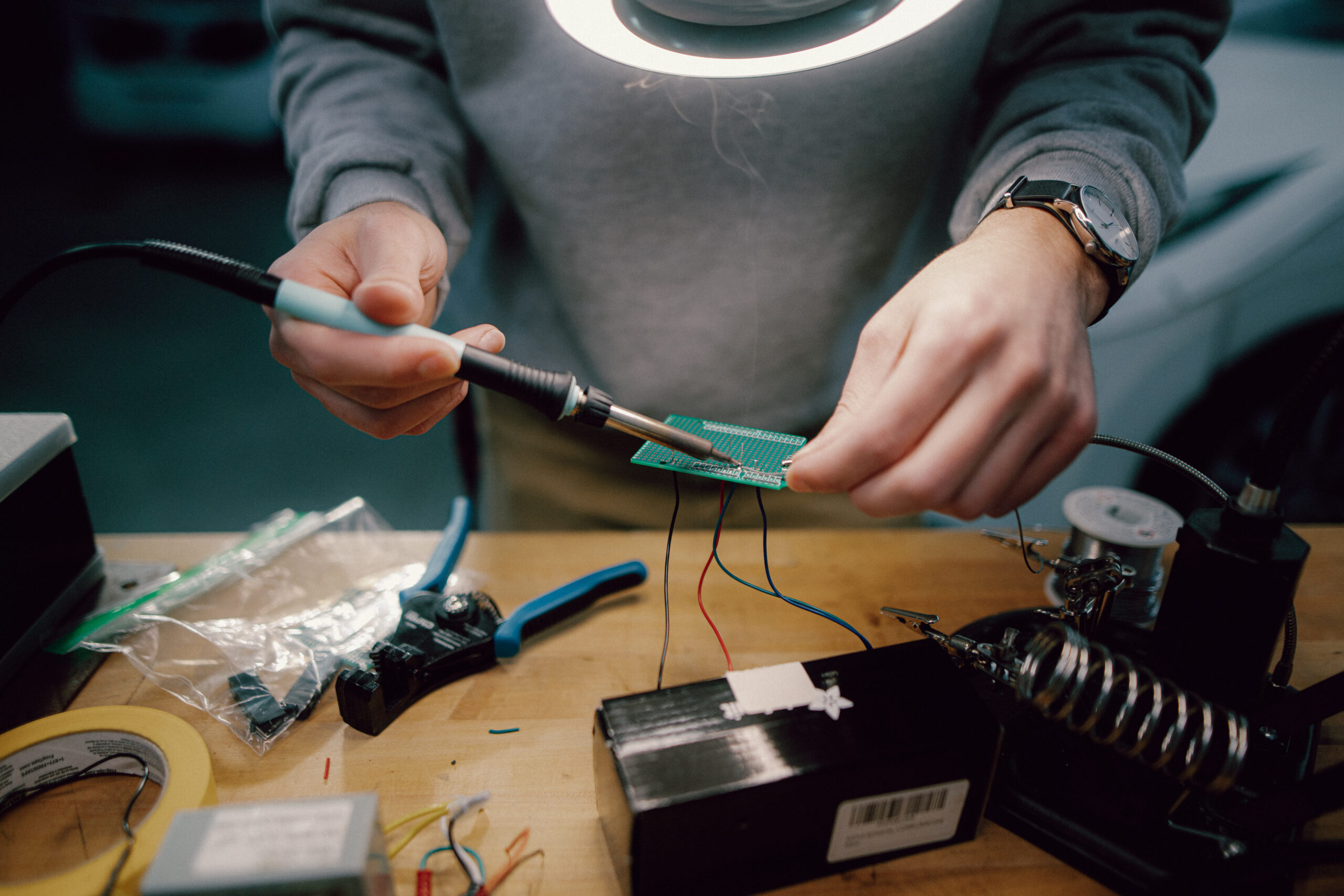 A close-up view of a student’s hands as they carefully work on an electronics project. The student is using a soldering iron to connect components on a circuit board. Various tools and materials, such as wire cutters, tape, and components, are spread out on the workbench. The focused light from a magnifying lamp highlights the intricate work being done, emphasizing the precision and skill involved in this hands-on learning experience.