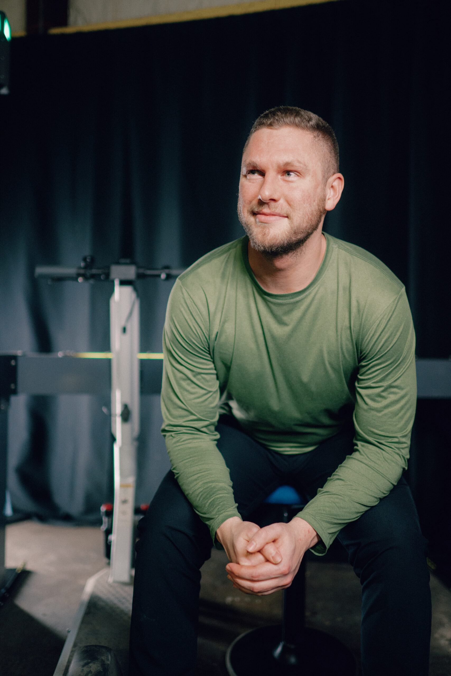 Peter Deppe, a 2020 Electrical Engineering graduate, is seated in a relaxed and confident pose, wearing a green long-sleeve shirt. The background features equipment, suggesting a technical or engineering environment.